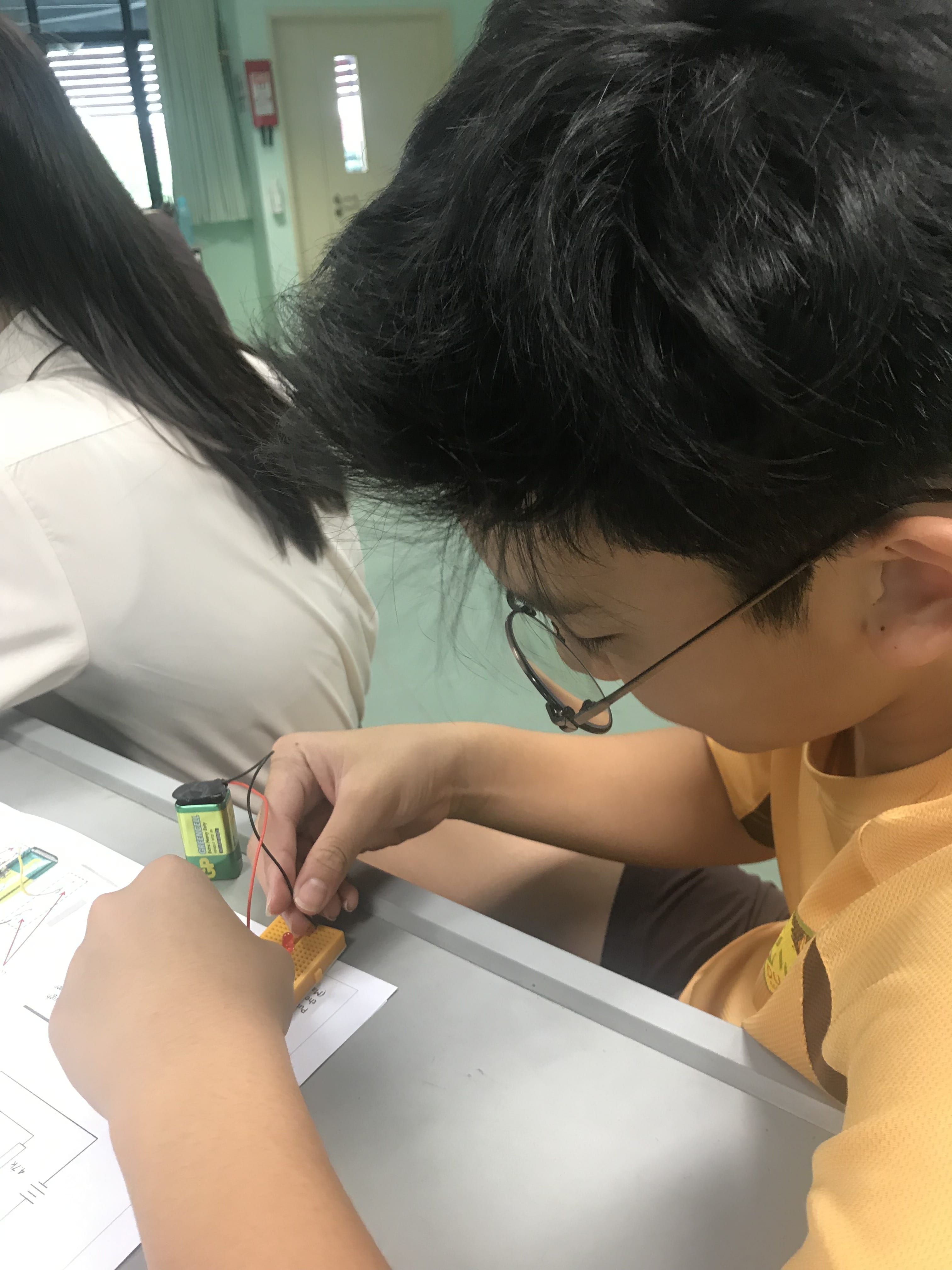 A young student with glasses leans over a desk, connecting wires to a battery and breadboard circuit.